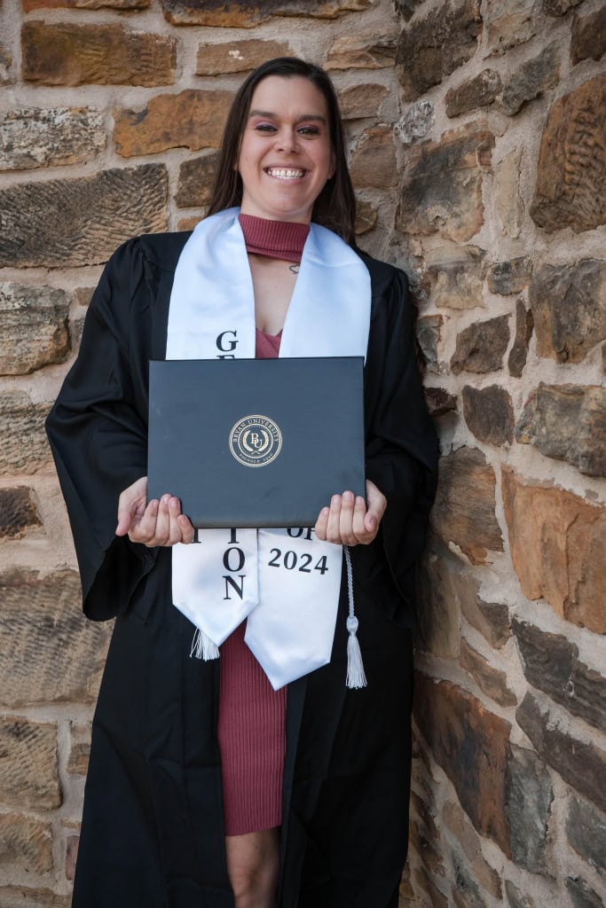 A smiling woman in a graduation gown holds a degree