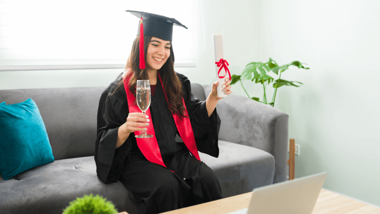 A woman in graduation regalia celebrating at home in front of a laptop