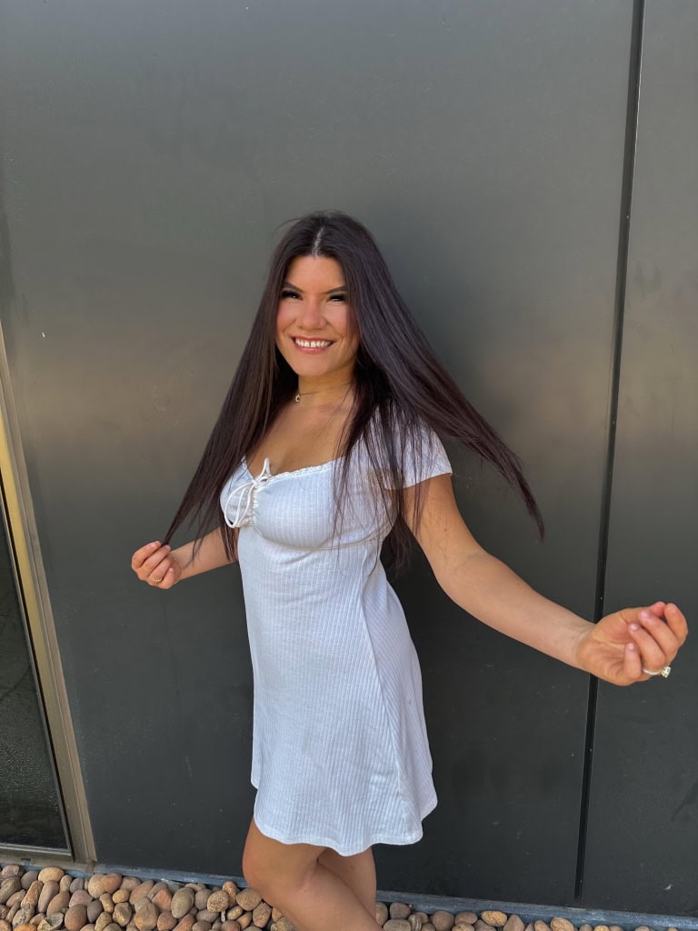 Bright young woman smiling outdoors, wearing a white dress, standing against a dark gray wall, representing online education and student success.